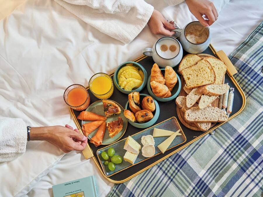 Ein Frühstückswagen mit Croissants, Käse, Trauben und einer Kaffeetasse auf einem Bett mit einer karierten Decke.