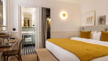 Habitación de hotel con cama, escritorio con silla, banca, baño con lavabo y espejo, y una lámpara de pared.