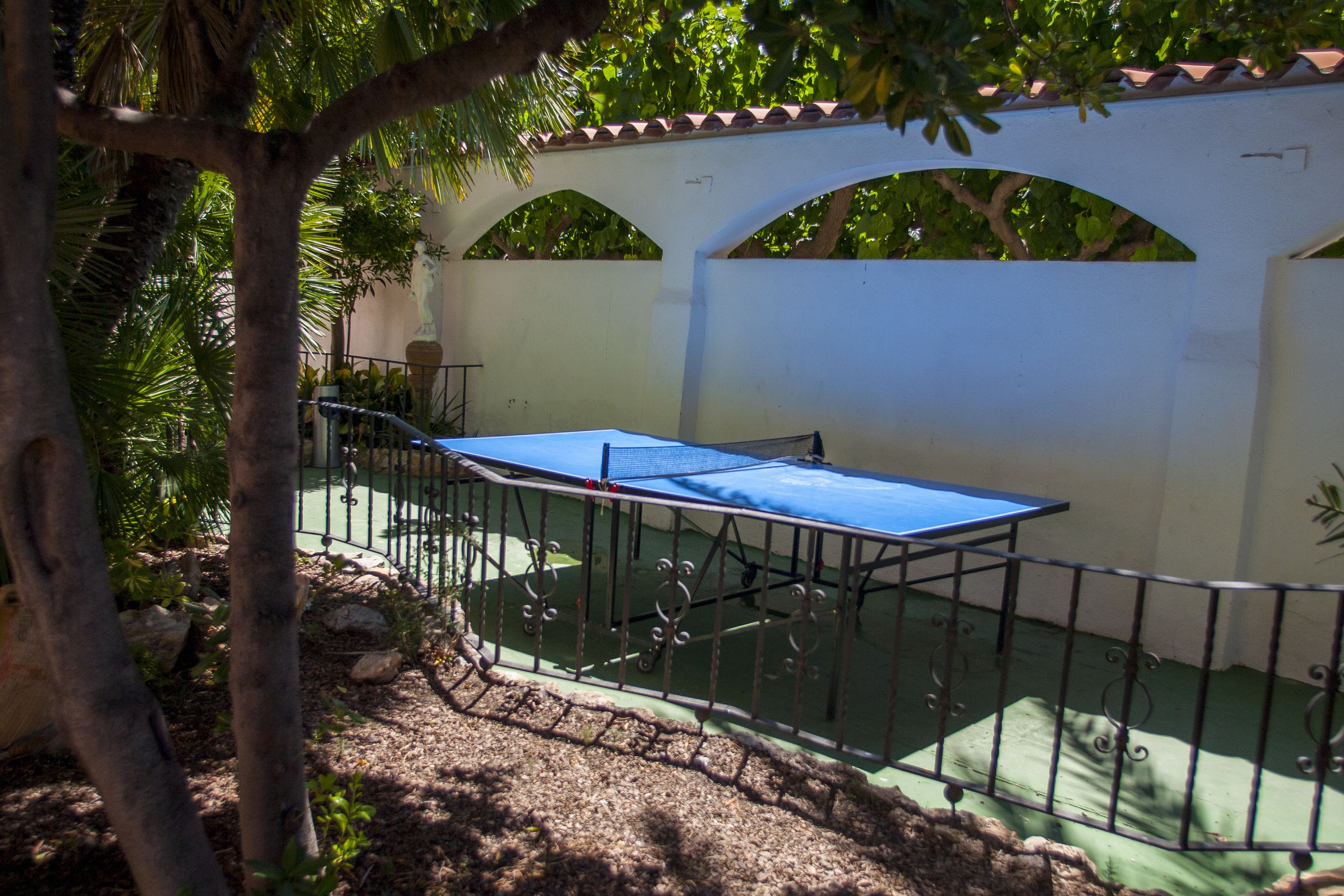 A courtyard with a table tennis table, surrounded by a metal fence, and a white wall with arches.