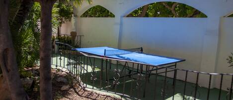 A courtyard with a table tennis table, surrounded by a metal fence, and a white wall with arches.