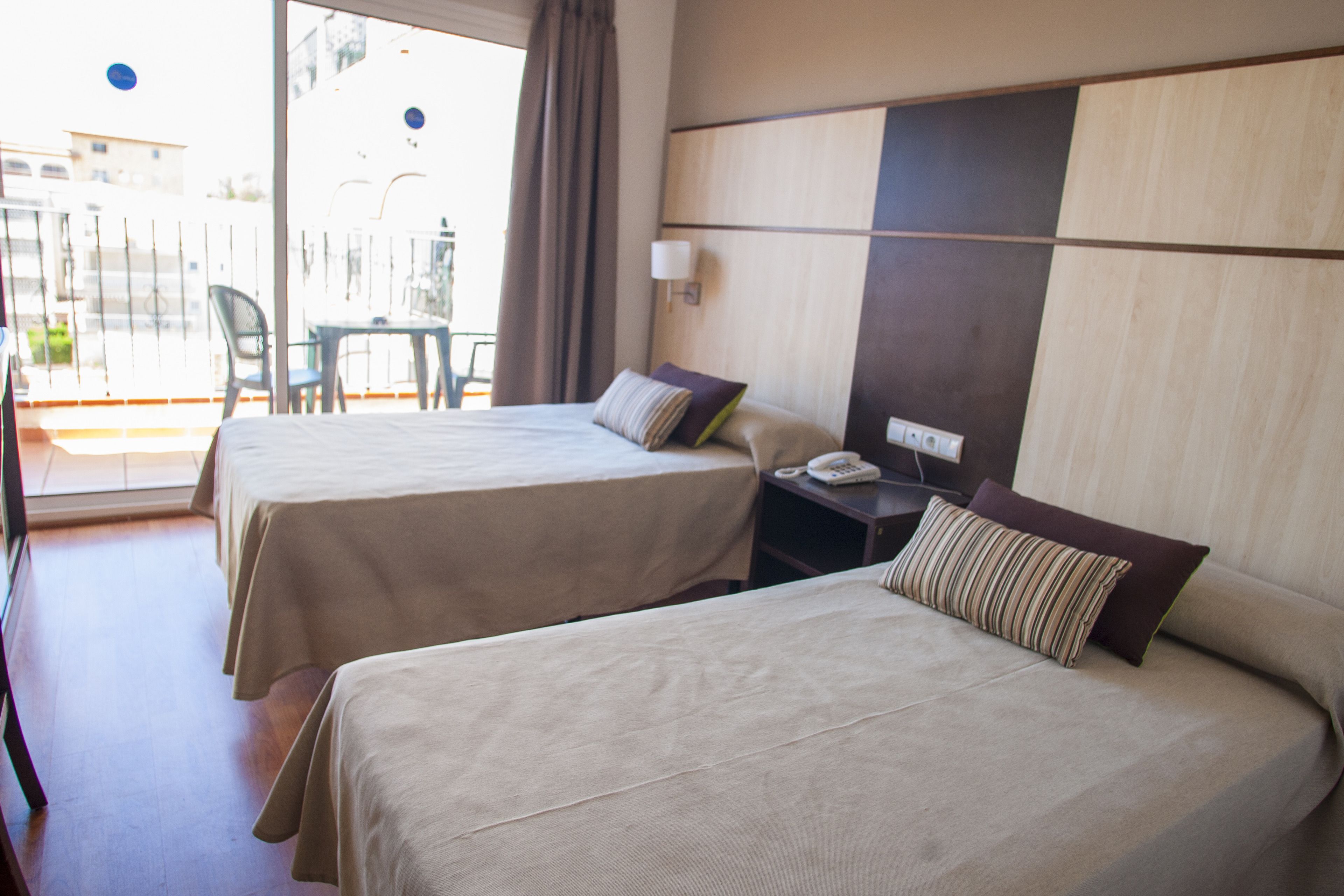 A hotel room with two beds, a small table, a chair, and a balcony with a view.