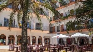 Un patio al aire libre con mesas y sillas, rodeado por un edificio con balcones y ventanas arqueadas.