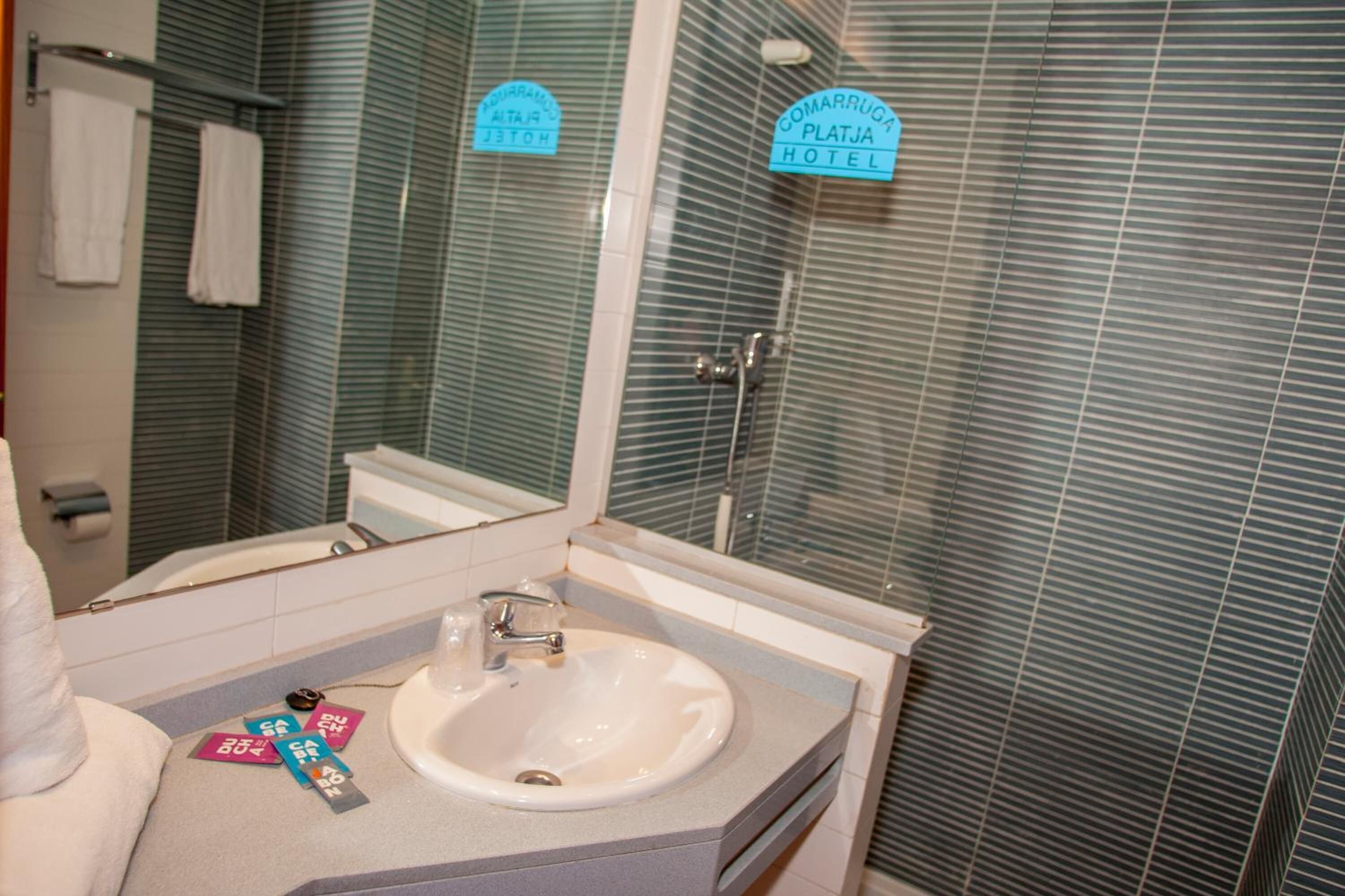 Un baño con lavamanos, espejo y perchero. Se observan señales con el nombre del hotel.