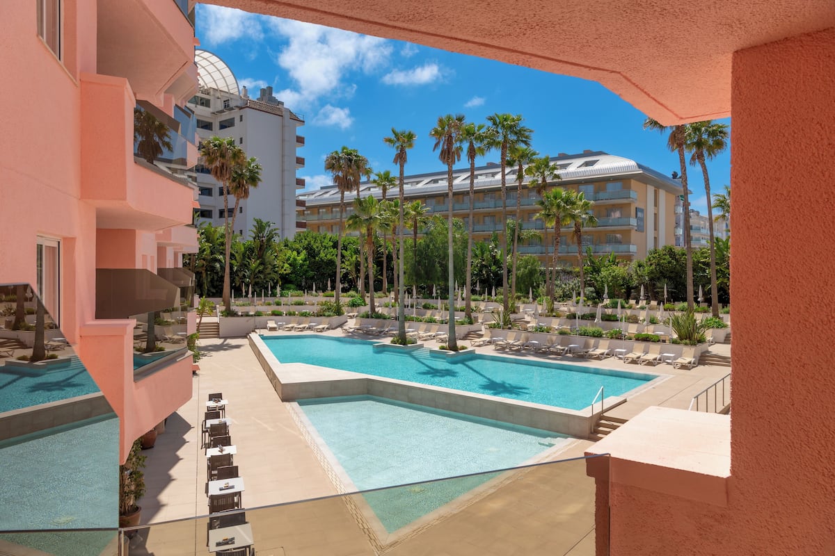 Un área de piscina hotelera con varias piscinas, palmeras y sillones de descanso.