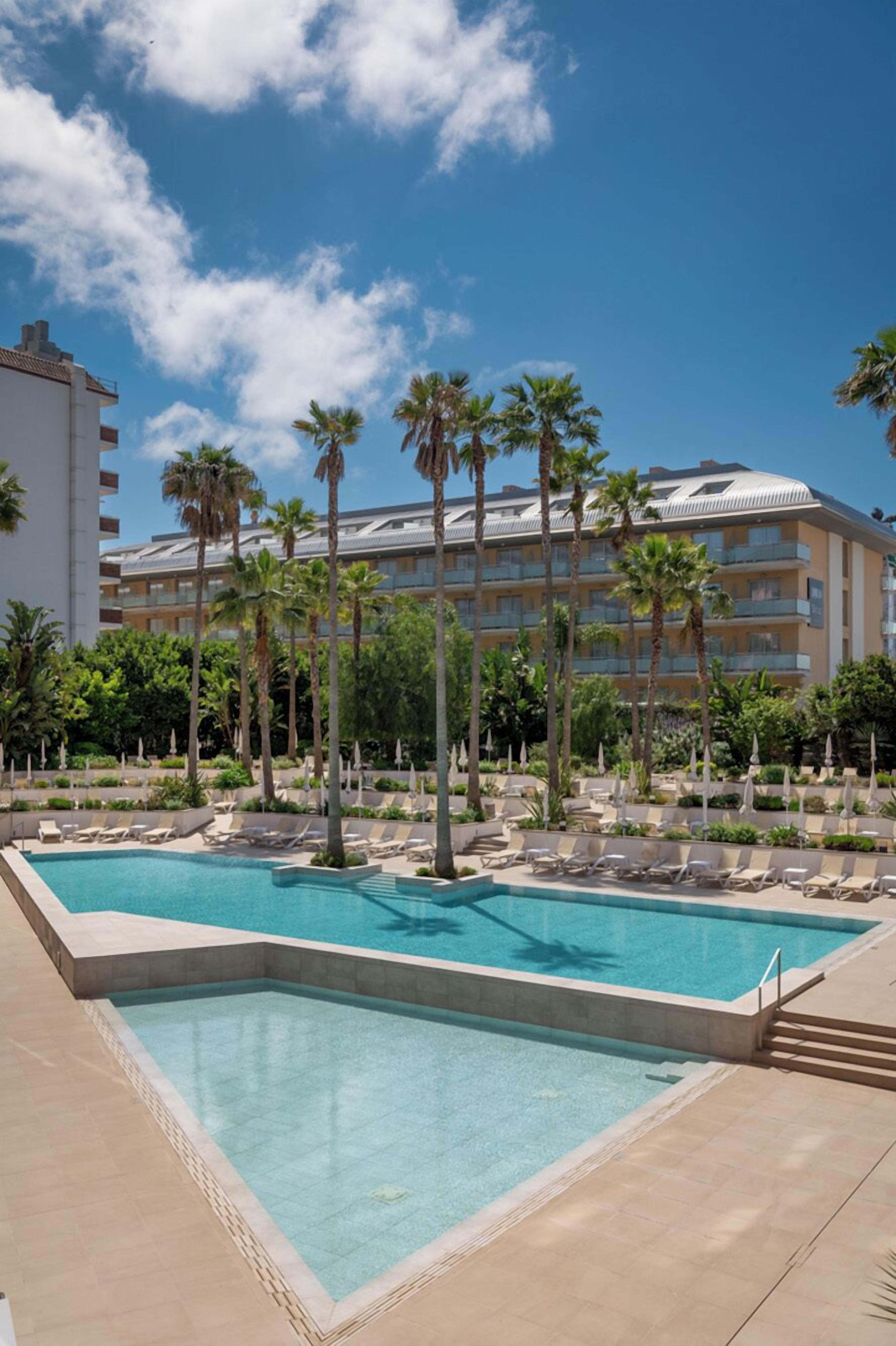 A swimming pool with lounge chairs, palm trees, and a hotel building in the background.