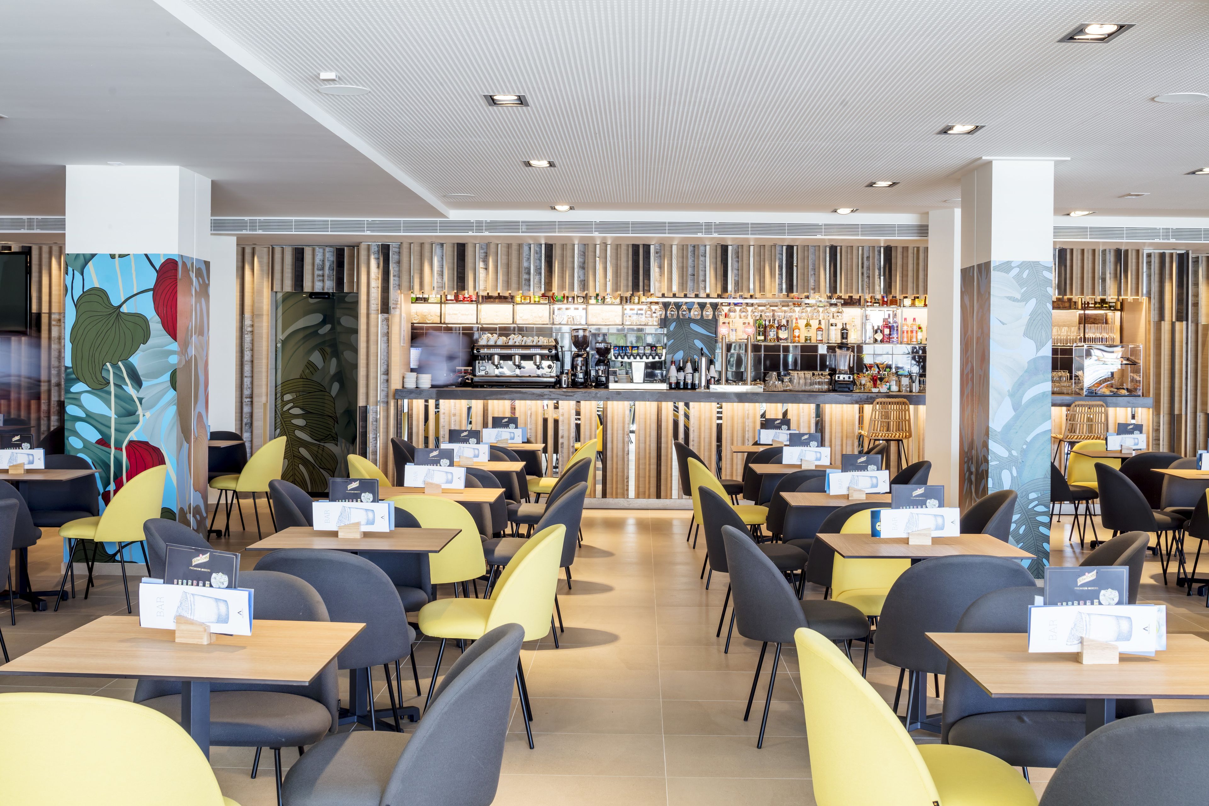 Un restaurante moderno con área de bar, con sillas amarillas y grises, mesas de madera y una pared decorativa con motivos de hojas.