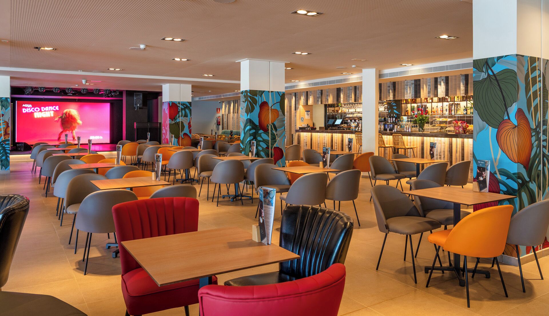 Un comedor moderno con varias mesas y sillas, una zona de bar con una variedad de botellas y un escenario con una pantalla grande mostrando un evento.