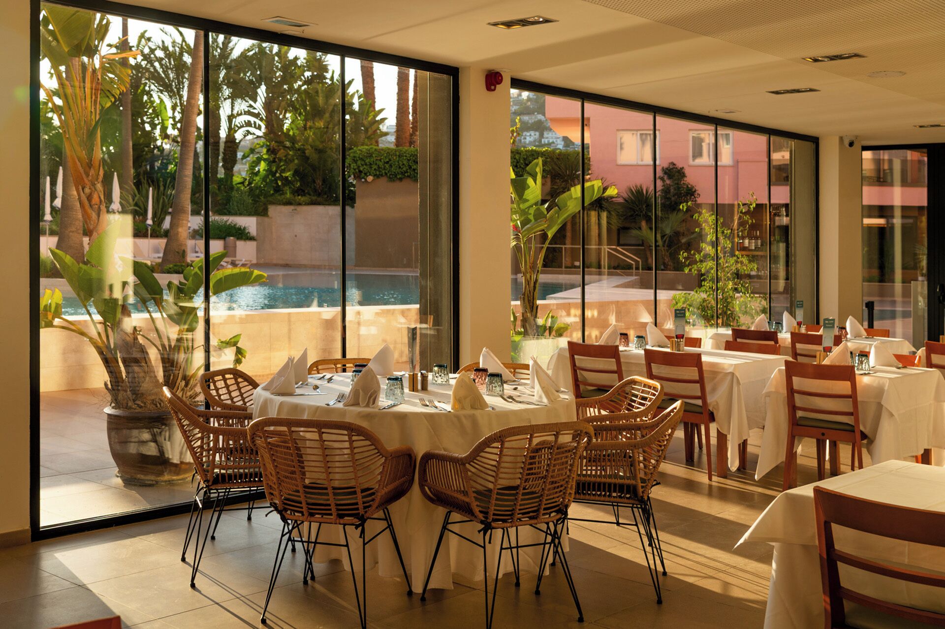 A restaurant with tables set for dining, wicker chairs, and a view of a pool and greenery outside.