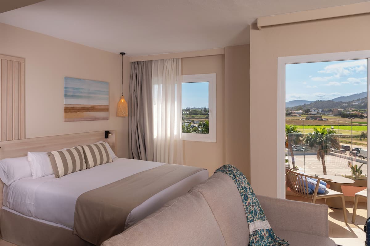 Una habitación de hotel con una cama grande, vistas al campo y un balcón con mobiliario exterior.