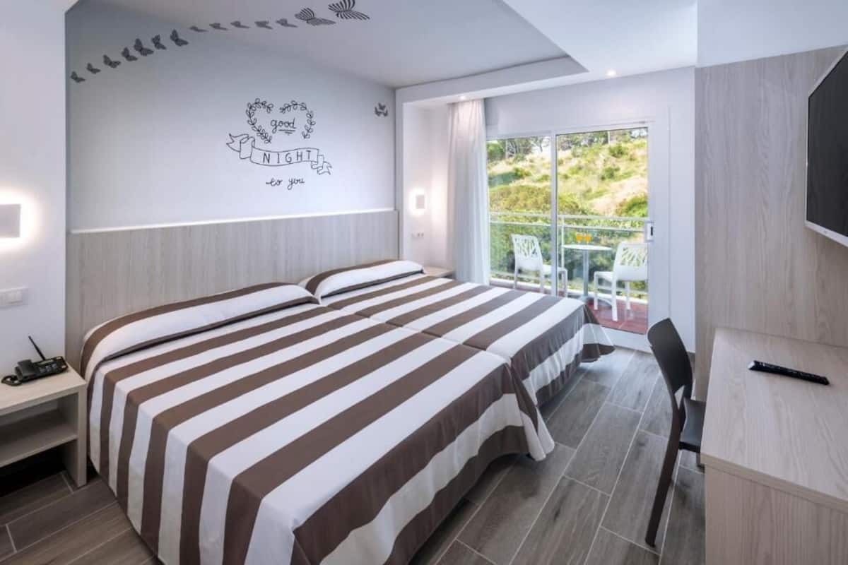 A hotel room with a large bed, a desk, a chair, a TV, and a balcony with outdoor seating.
