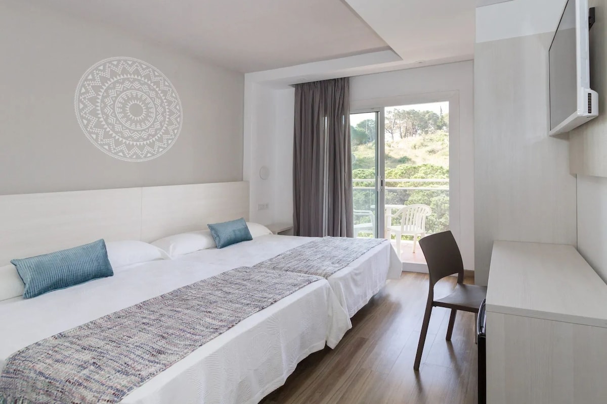 A bedroom with a bed, a desk, a chair, a large window, and a wall decoration.
