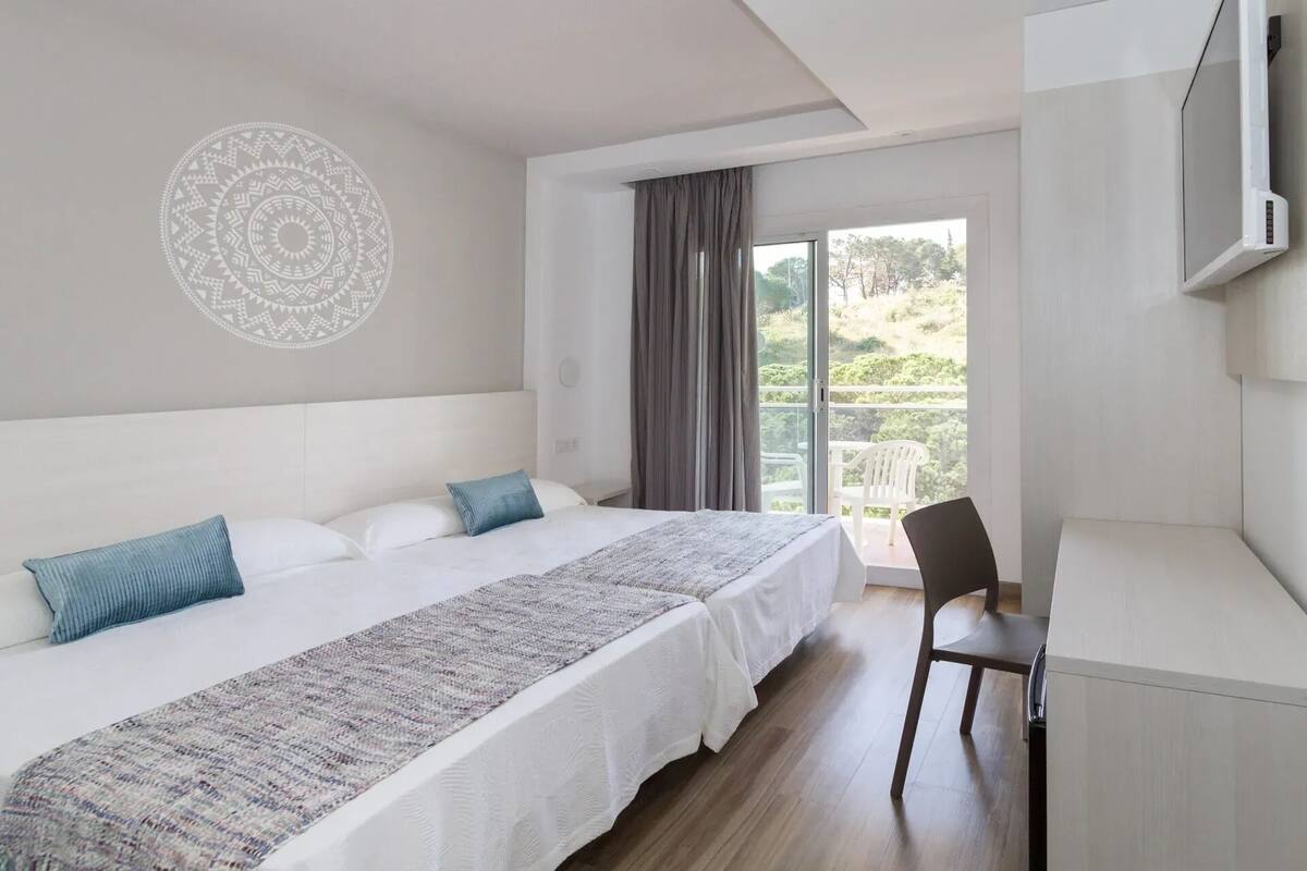 A bedroom with a bed, a desk, a chair, a large window, and a wall decoration.