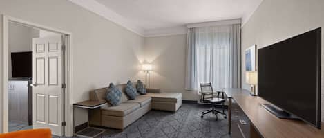 A hotel room with a sectional sofa, a TV, a desk, and a chair.