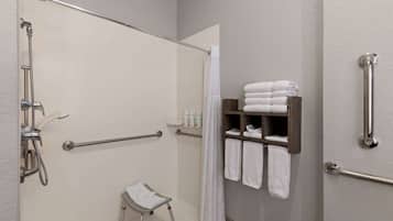A bathroom with a shower, grab bars, a small stool, and a shelf with towels.