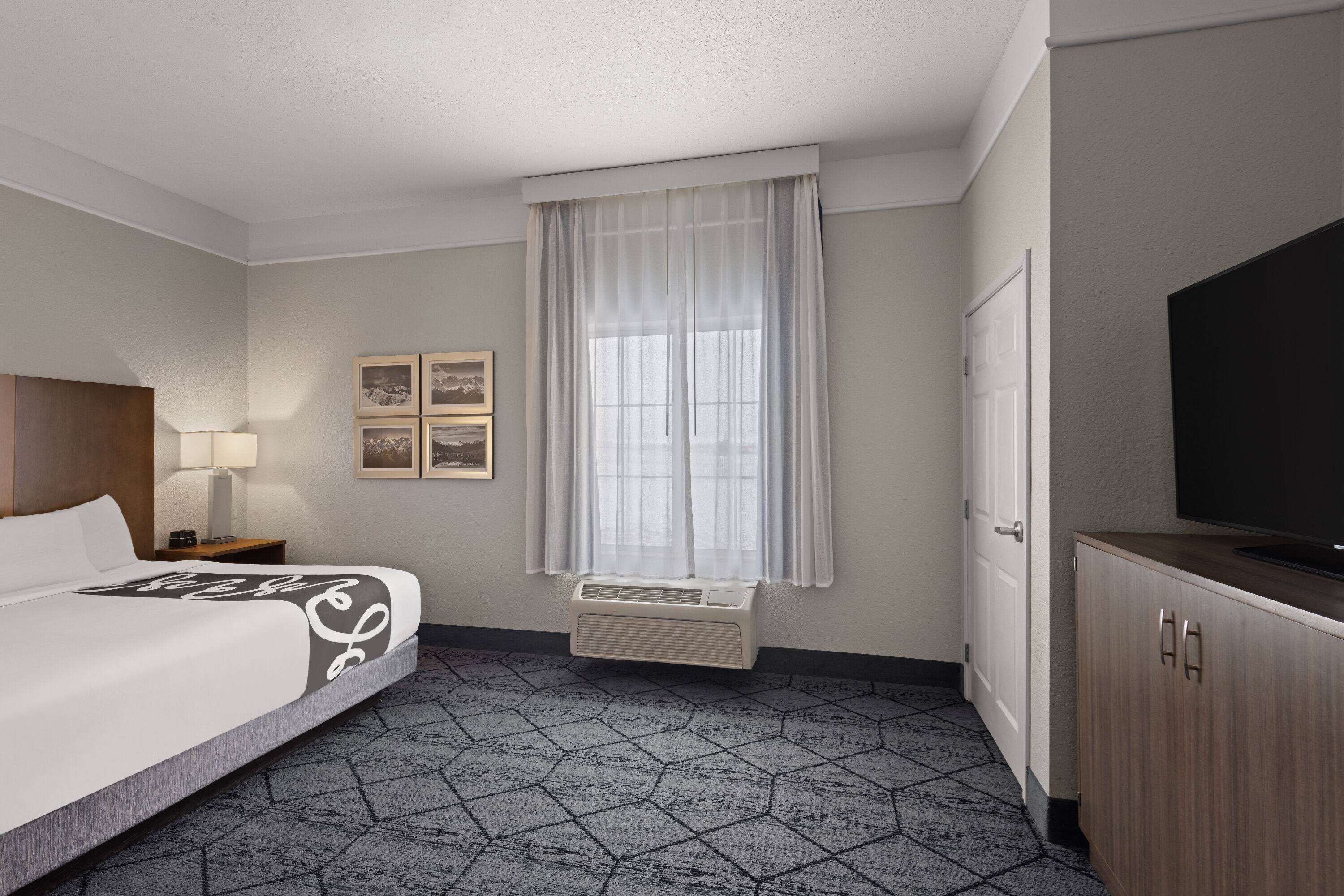 Habitación de hotel con cama, televisor, ventana con cortinas, aire acondicionado de pared y cuadros enmarcados.