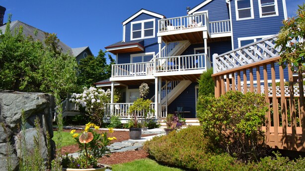 A two-story house with a balcony, a stone wall, and a garden with flowers and shrubs. - The Heron Inn & Day Spa (La Conner)