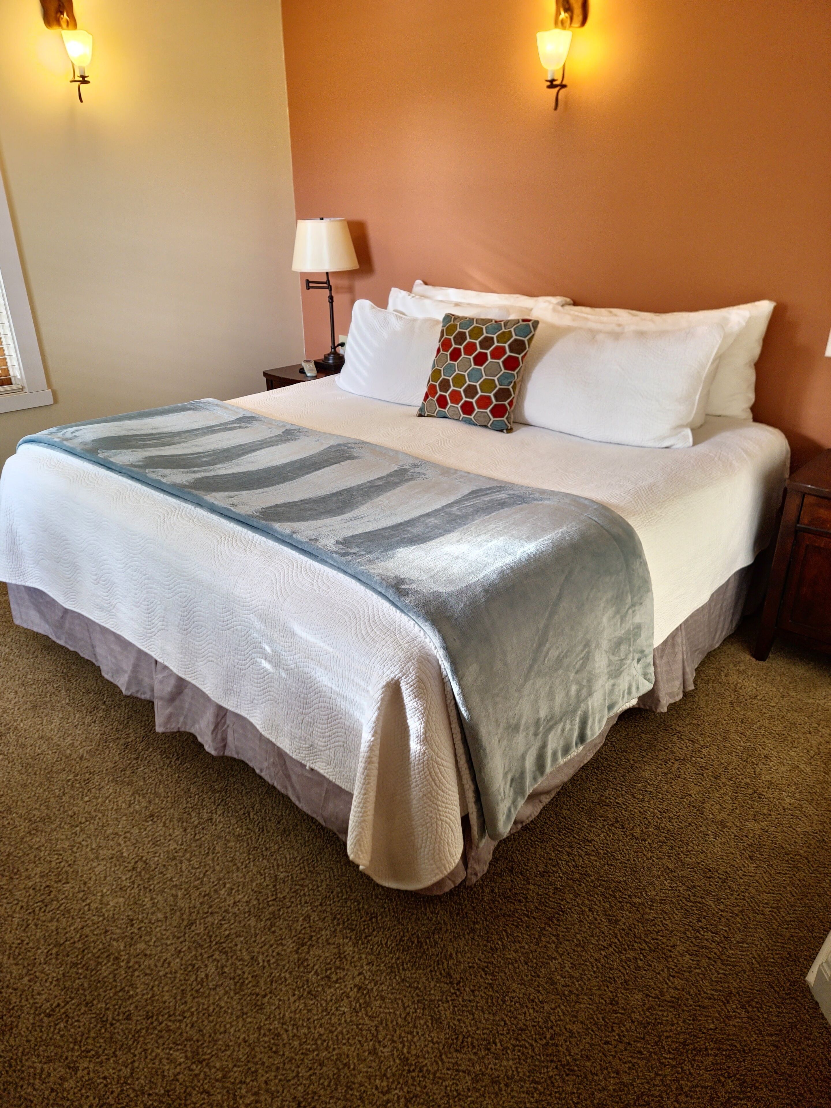 Una cama bien tendida con una manta estampada, almohadas blancas y una almohada decorativa. Hay luces fijadas a la pared y una mesita de noche con una lámpara.