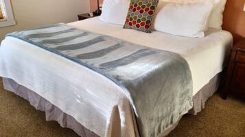 Una cama bien tendida con una manta estampada, almohadas blancas y una almohada decorativa. Hay luces fijadas a la pared y una mesita de noche con una lámpara.