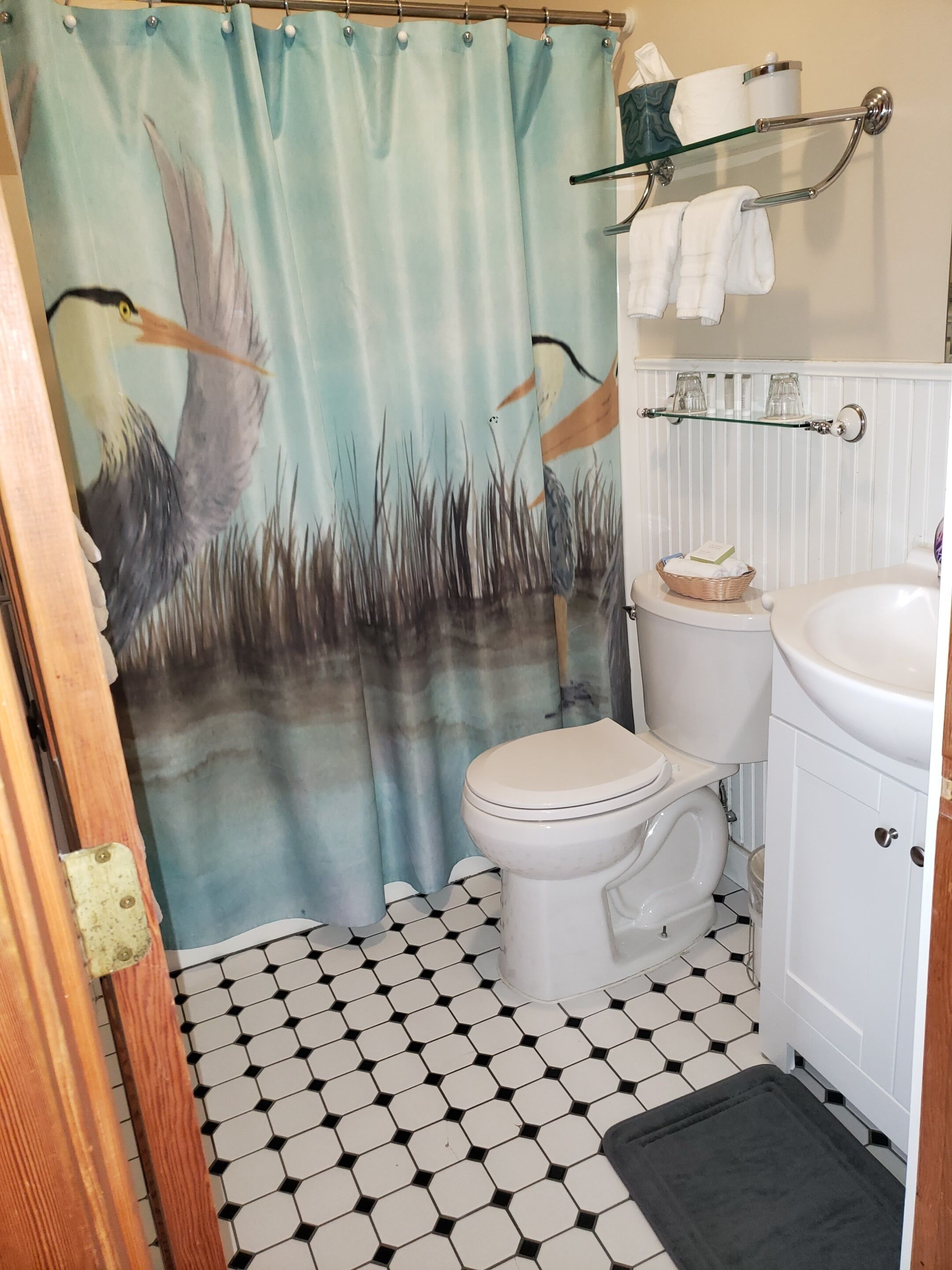 A bathroom with a toilet, a sink, a glass shelf with towels, a shower curtain with a bird and grass design, and a hexagonal tiled floor.