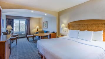 A hotel room with a large bed, a desk, a chair, and a small table. There is a window with curtains, a picture on the wall, and a lamp on the desk.