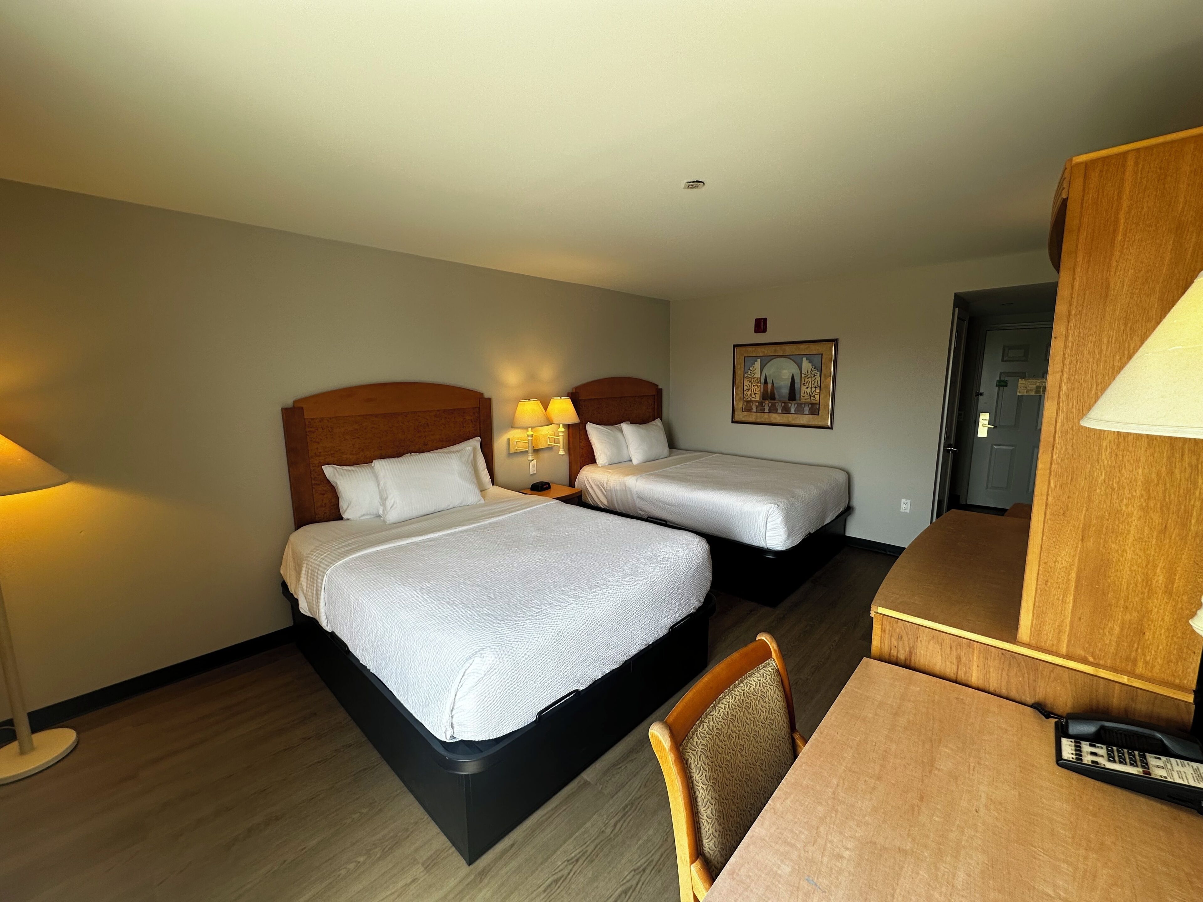 Quarto de hotel com duas camas, uma escrivaninha, uma cadeira e uma luminária.