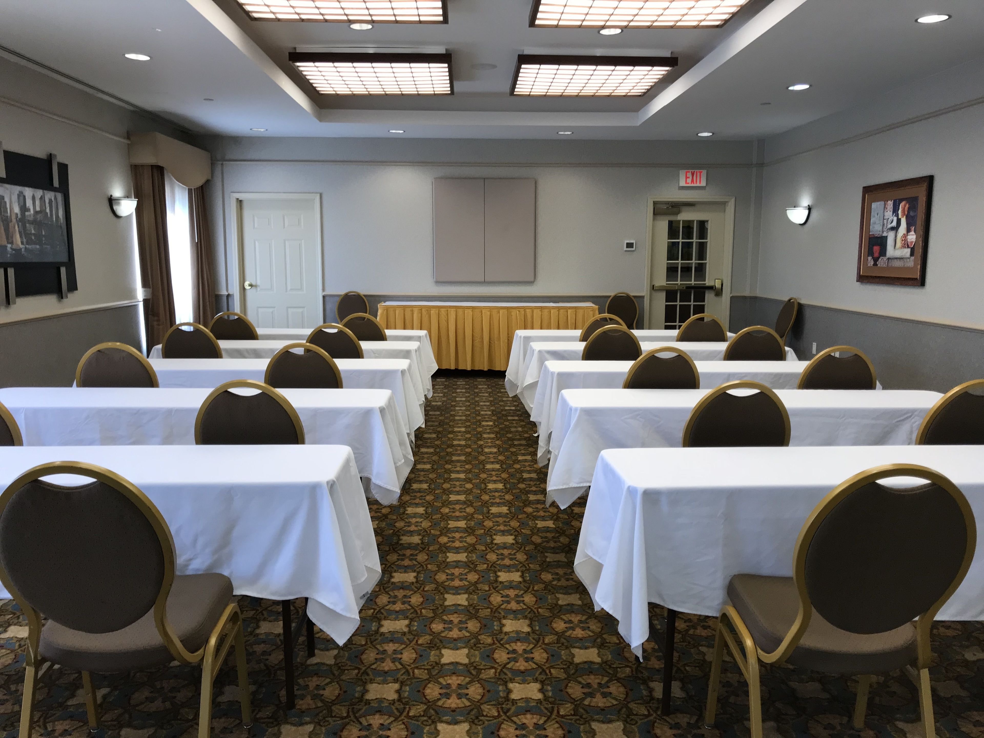 A conference room with rows of tables and chairs, a carpeted floor, and a door with an exit sign.