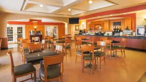 A spacious restaurant with multiple dining tables and chairs, a kitchen area, and a large window. - La Quinta Inn & Suites by Wyndham Islip - MacArthur Airport (Bohemia)