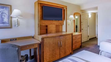 A hotel room with a wooden desk, a television mounted on a wooden panel, a bed with striped bedding, and a door leading to another room.