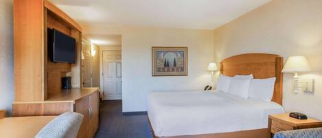 A hotel room with a large bed, a desk with a flat-screen TV, a chair, a bedside table with a lamp, and a framed picture on the wall.