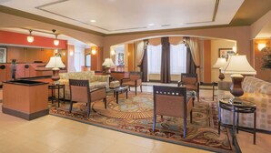 A hotel lobby with a reception desk, seating area with armchairs and sofas, patterned carpet, and ambient lighting. - La Quinta Inn & Suites by Wyndham Islip - MacArthur Airport (Bohemia)
