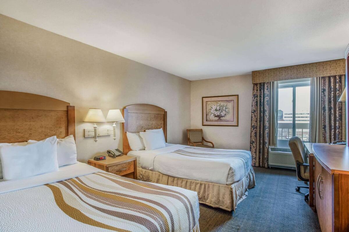Une chambre d’hôtel avec deux lits, des lampes de chevet, un bureau, une chaise, un tableau encadré au mur et une fenêtre avec des rideaux.