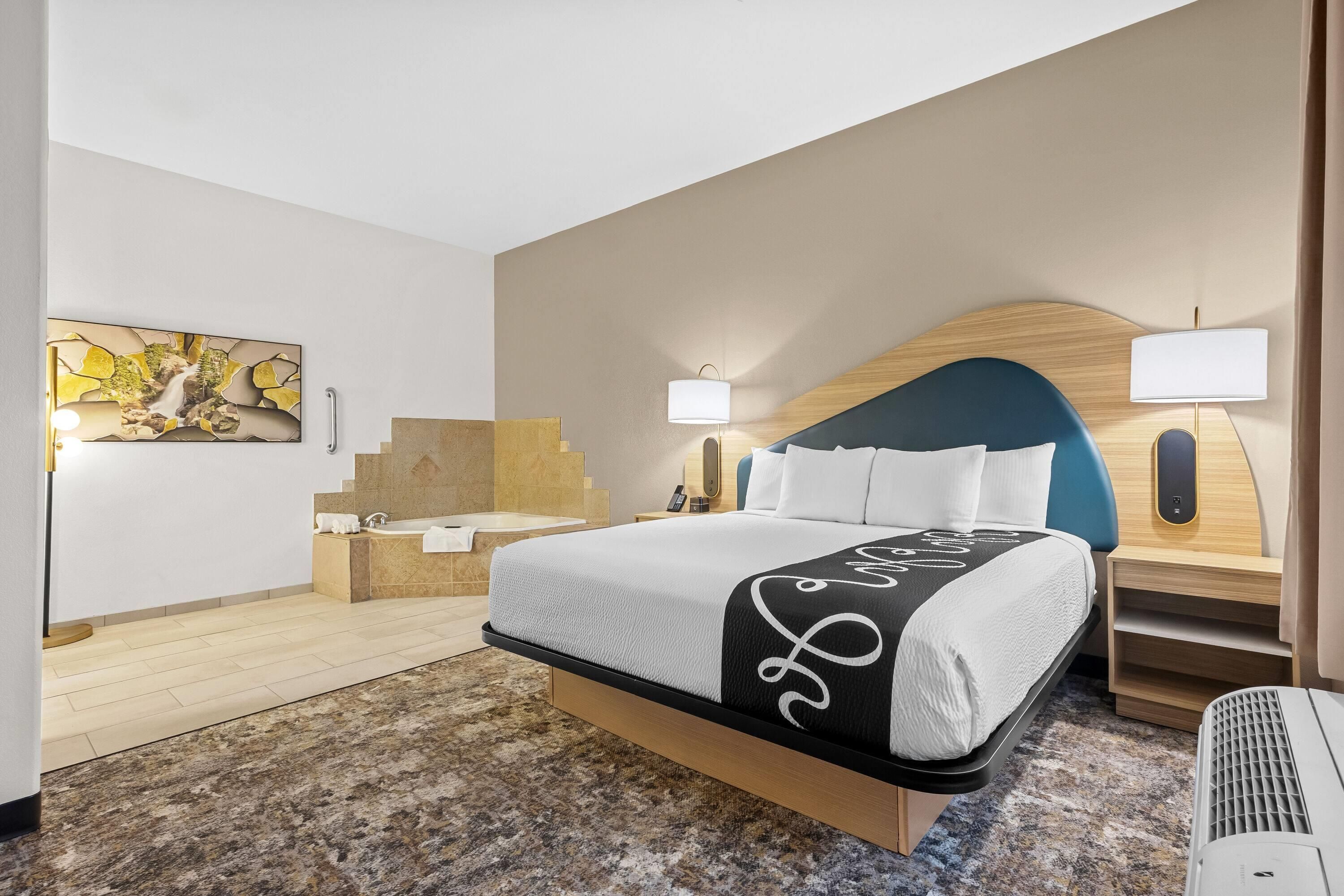 A hotel room with a bed, bedside tables, a headboard, a wall-mounted artwork, and a carpeted floor.