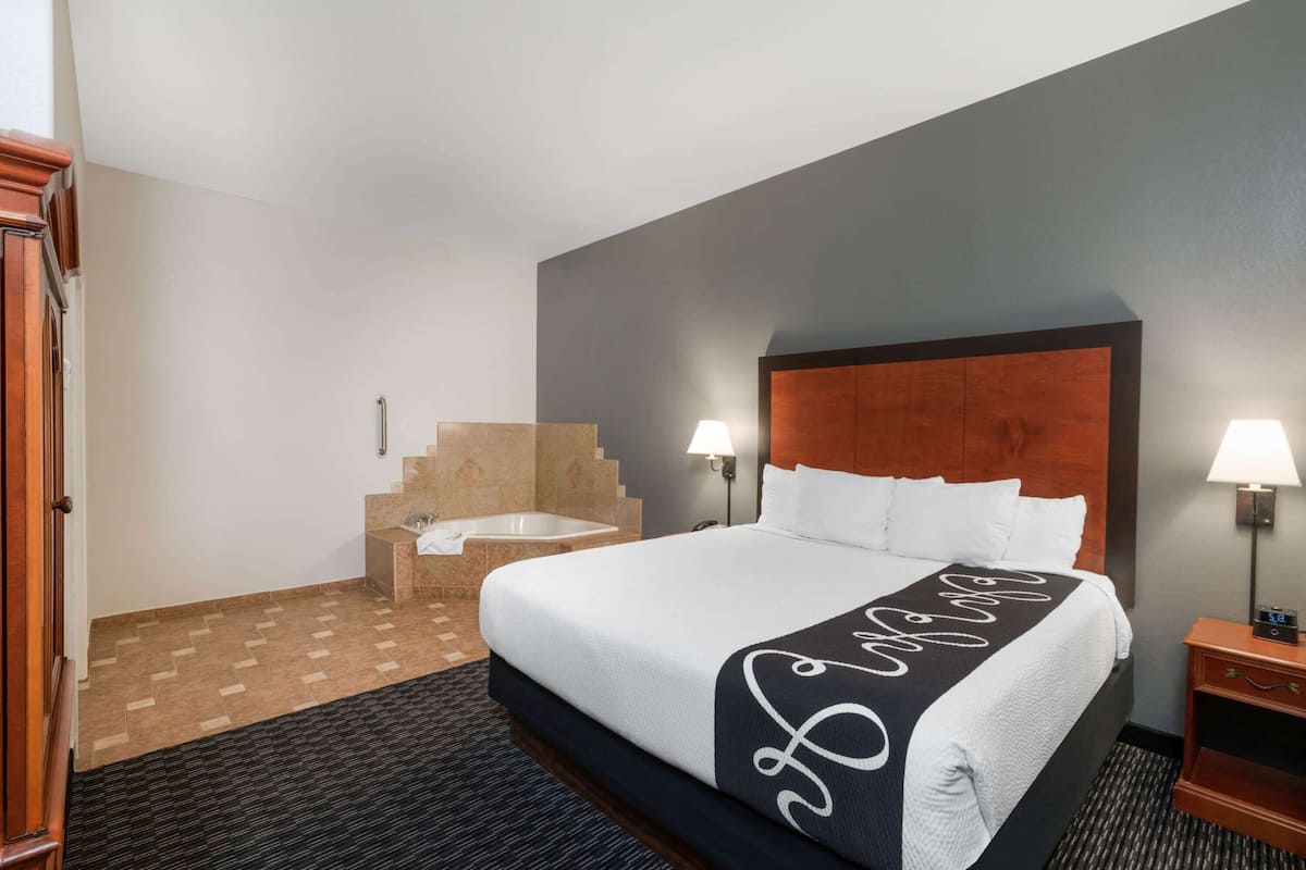A hotel room with a large bed, two bedside lamps, a wooden headboard, a bathroom with a bathtub, and a carpeted floor.