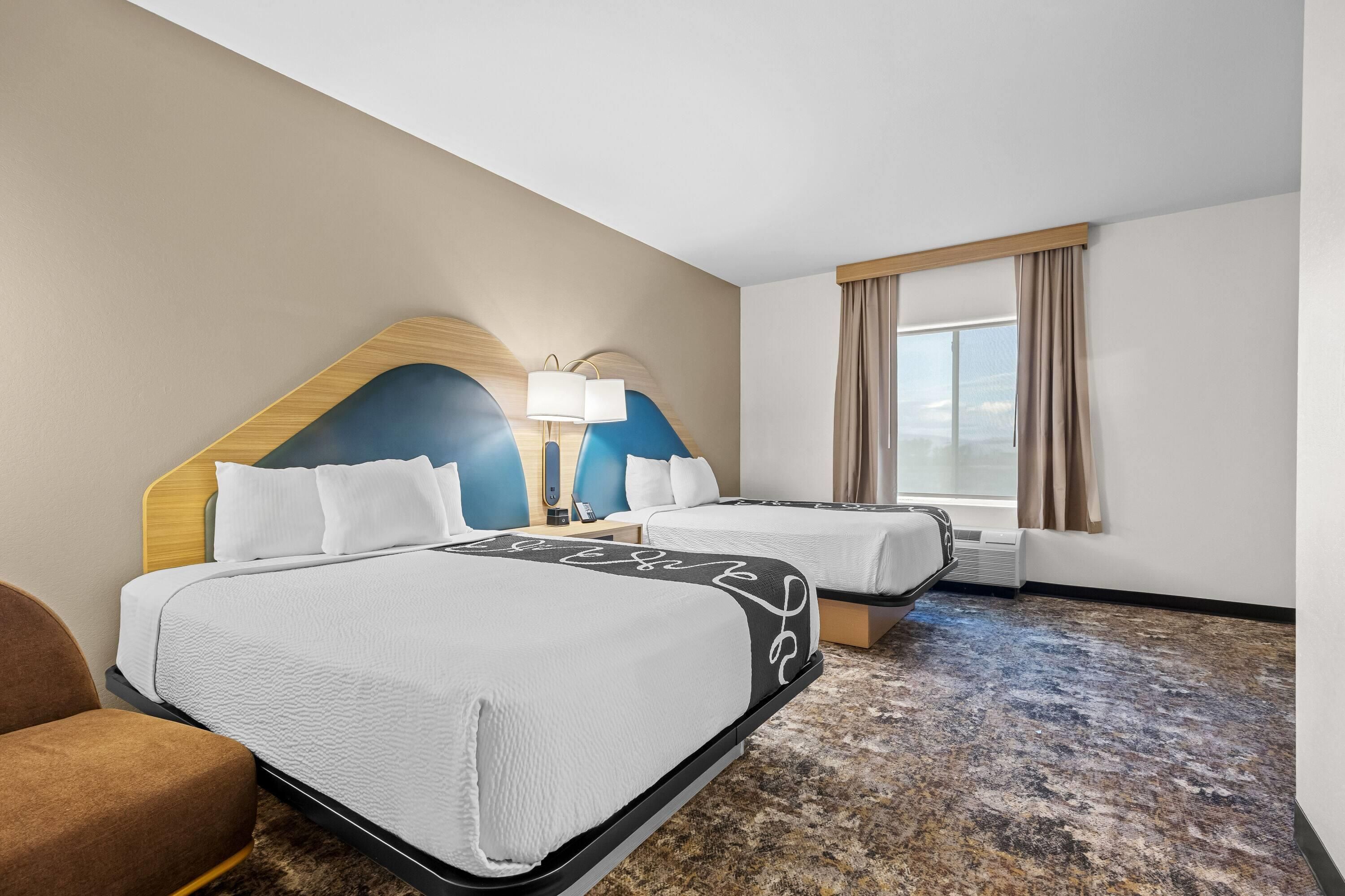 A hotel room with two beds, a headboard, a bedside table with a lamp, a window with curtains, and a carpeted floor.
