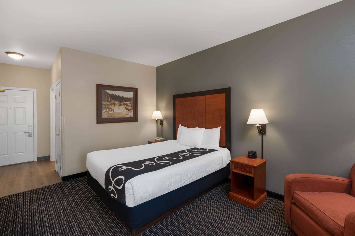 A hotel room with a large bed, two bedside lamps, a wooden nightstand, a framed picture on the wall, and a door to the left.
