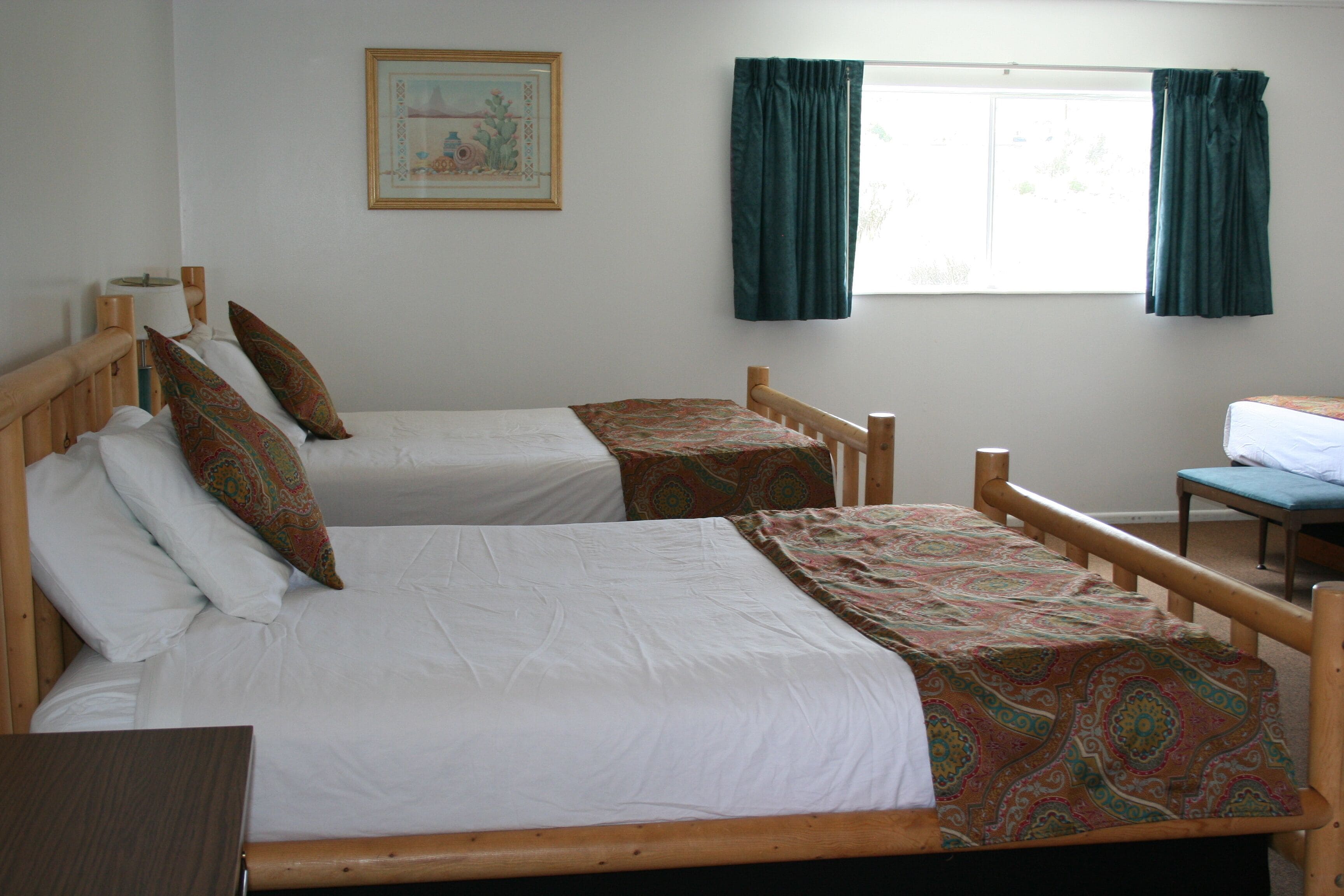 A room with two single beds, a wooden headboard, a bedside table, a window with curtains, and a framed picture on the wall.