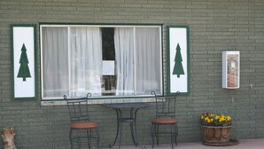 Un muro de ladrillo con una ventana, dos sillas de metal, una mesita y una maceta con flores.