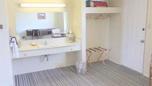 Habitación de hotel con un tocador blanco, un espejo, un lavamanos, una mesita con una taza, un perchero y una alfombra estampada.