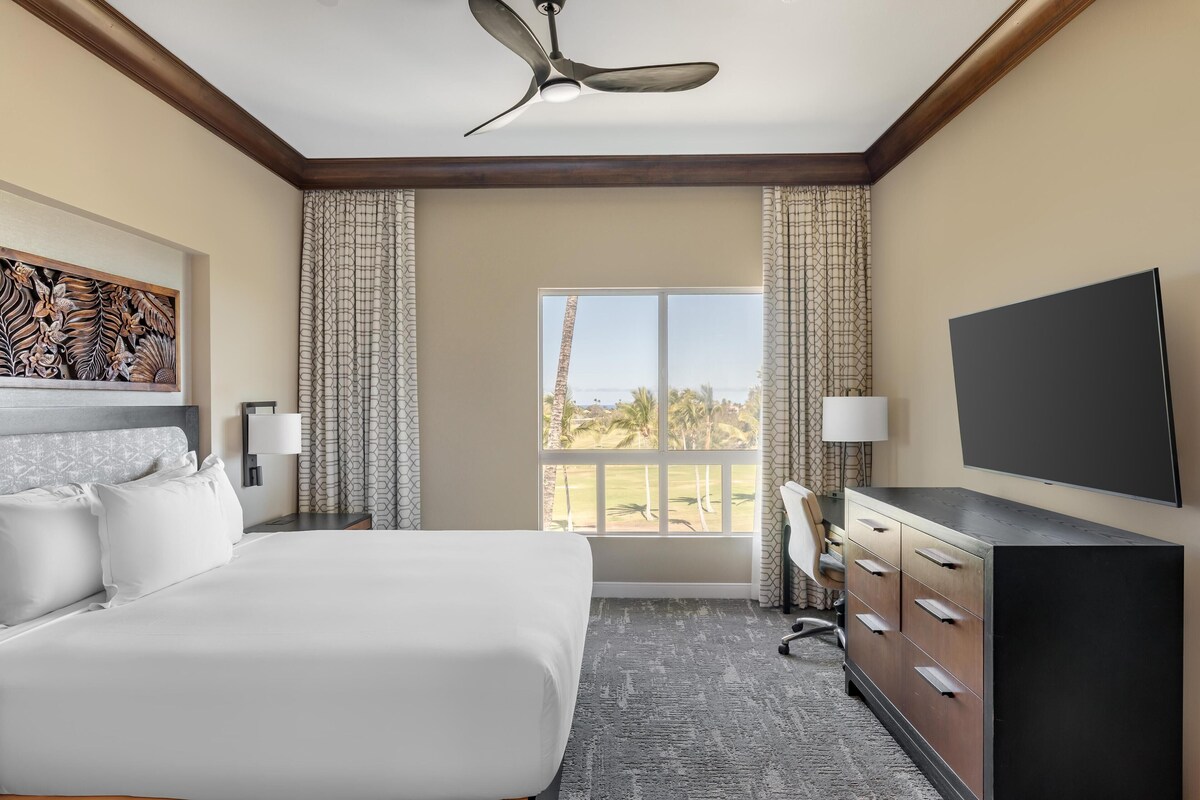 Een hotelkamer met een groot bed, een bureau, een televisie en een raam met uitzicht op palmbomen.