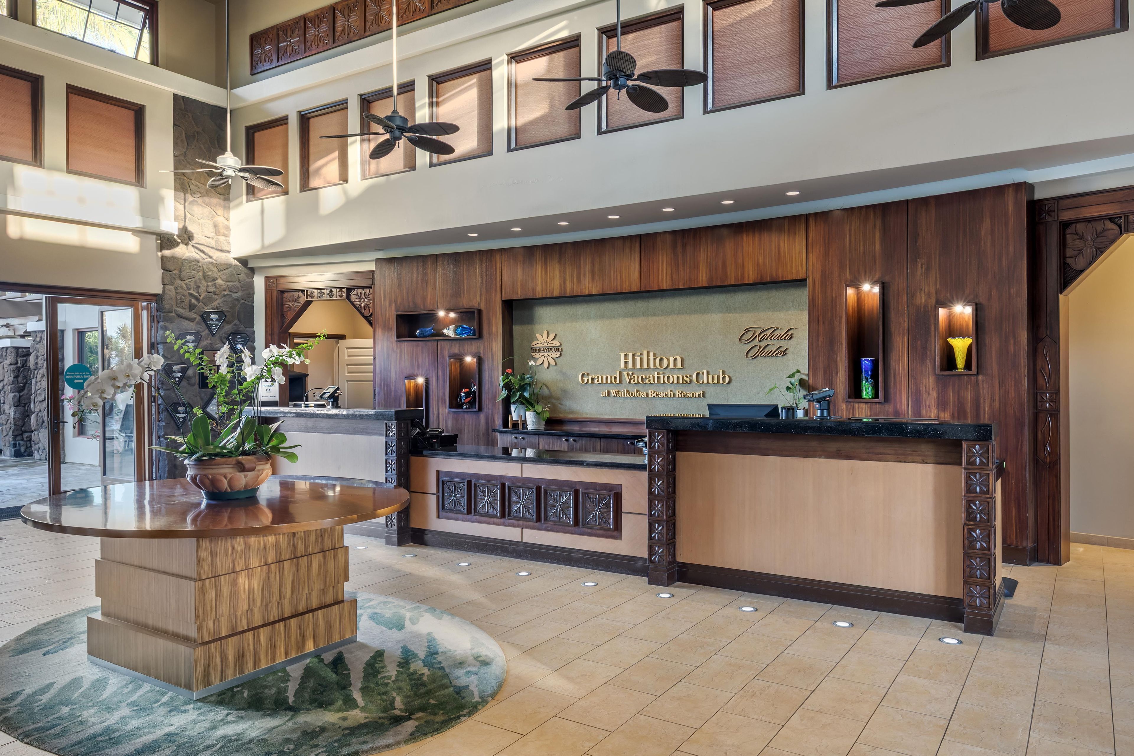 A hotel reception area with a wooden reception desk, a large sign for Hilton Grand Vacations Club, and multiple ceiling fans.