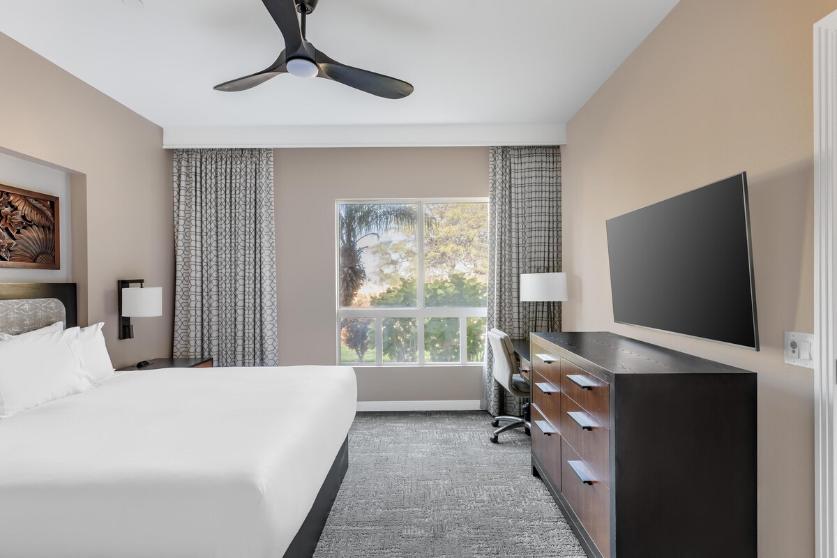 Hotelkamer met een bed, bureau, televisie, plafondventilator en een raam met gordijnen.