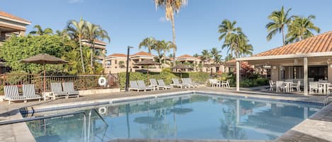 Una piscina con sillas de descanso y sombrillas, rodeada de palmeras y edificios con techos de tejas.