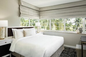 Une chambre d’hôtel avec un grand lit, une table de chevet, une lampe et une fenêtre donnant sur des palmiers.