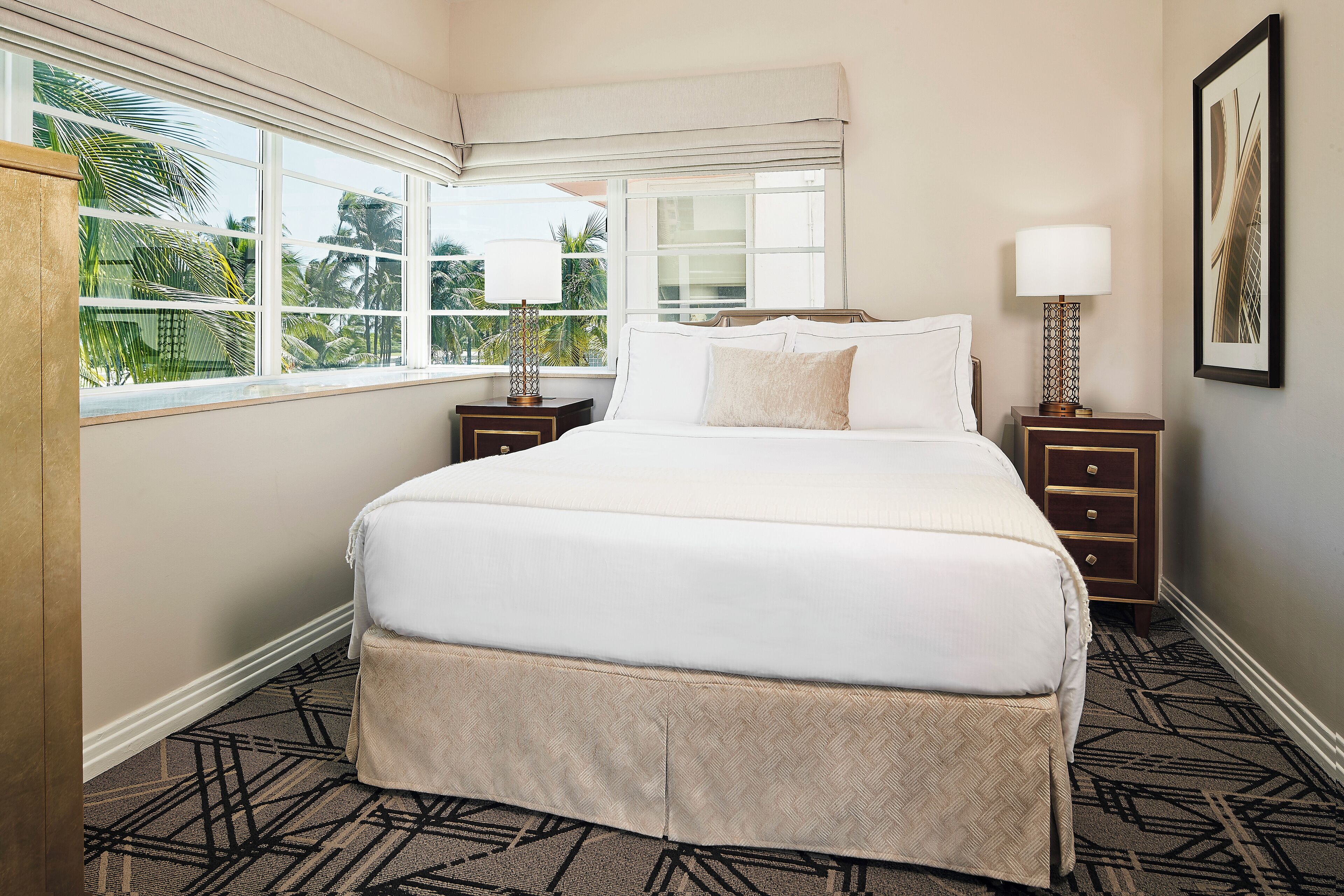 A neatly made bed with a white comforter and pillows, a bedside table with a lamp, and a window with a view of palm trees.