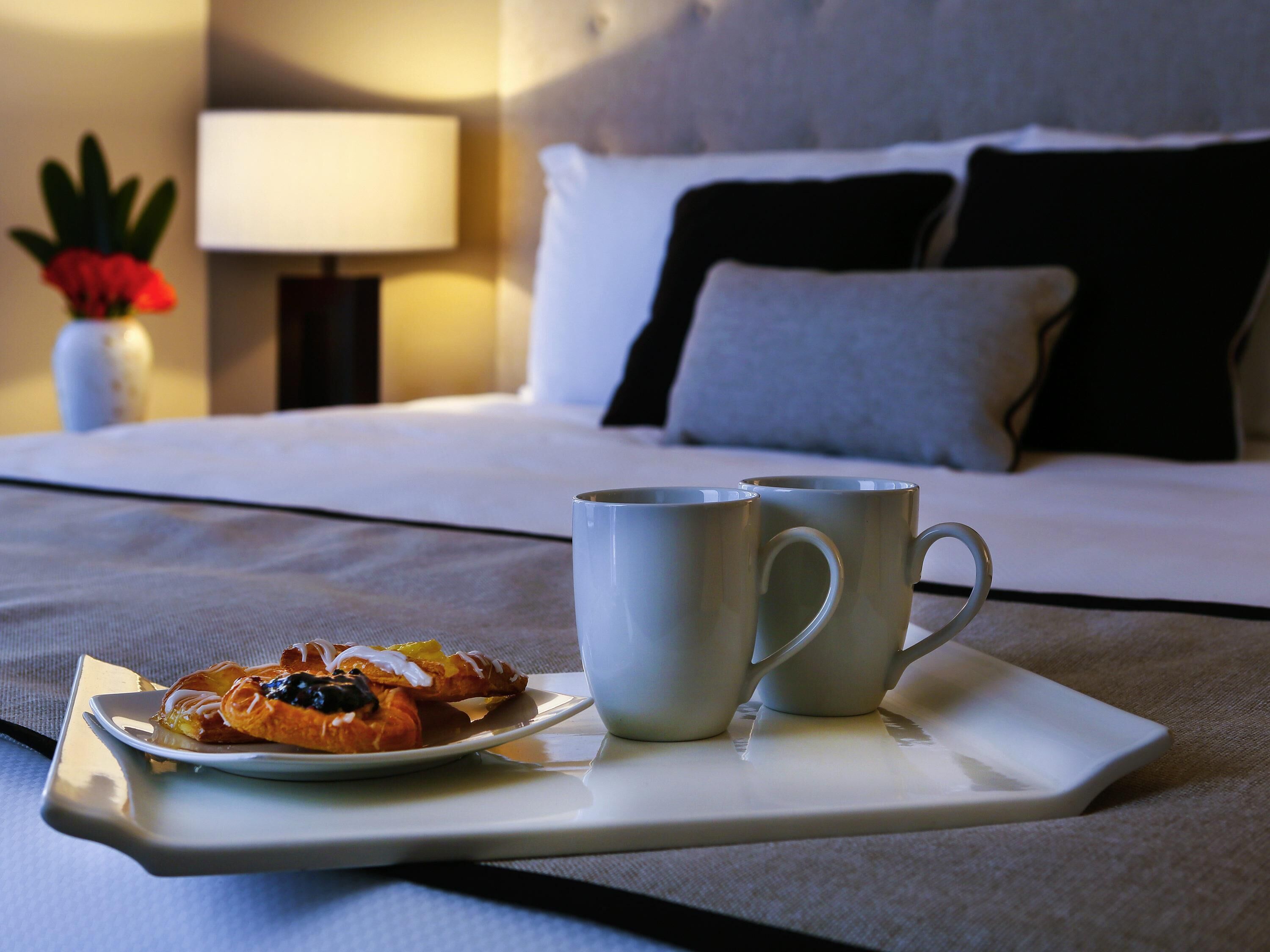 A neatly made bed with pillows, a bedside table with a lamp, and a tray with a pastry and two mugs on a table.