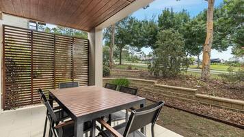 Eine überdachte Terrasse mit einem Holztisch und Stühlen, mit Blick auf Bäume und einen Park im Hintergrund.