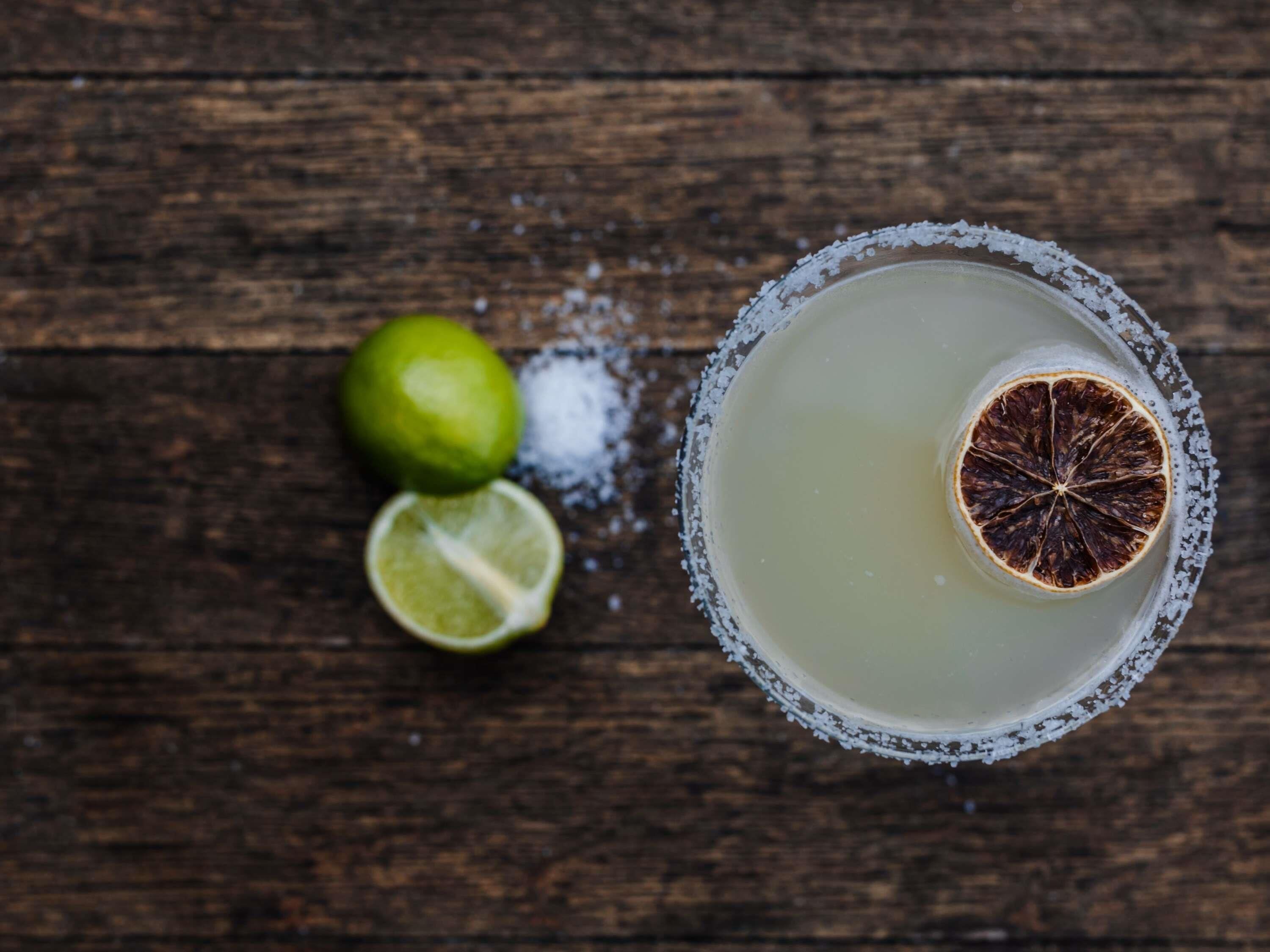 A margarita glass with a salted rim and a dried lime slice, a whole lime, and a half lime on a wooden surface.