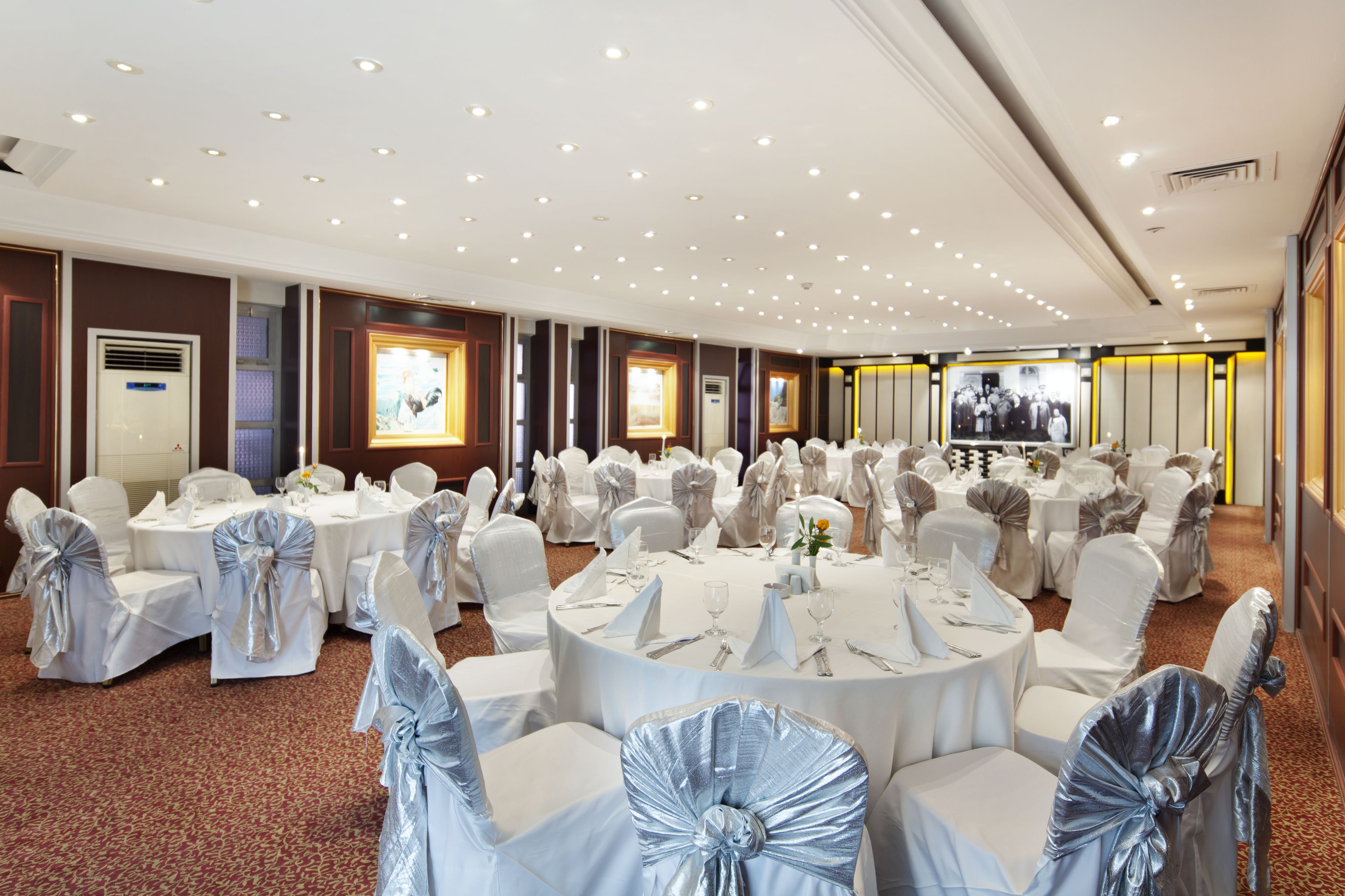 A banquet hall with round tables, white chairs with blue sashes, and a carpeted floor.
