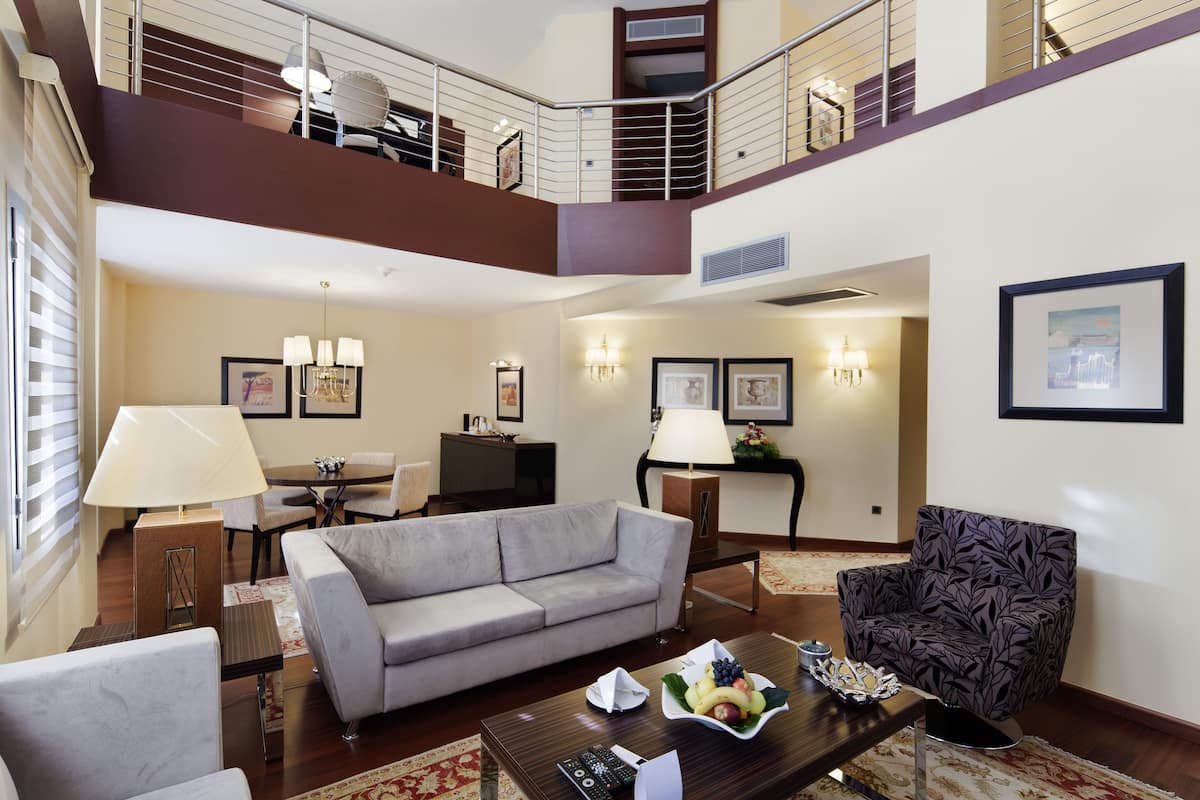 A spacious living area with a grey sofa, a patterned armchair, and a coffee table with remote controls and a fruit bowl.
