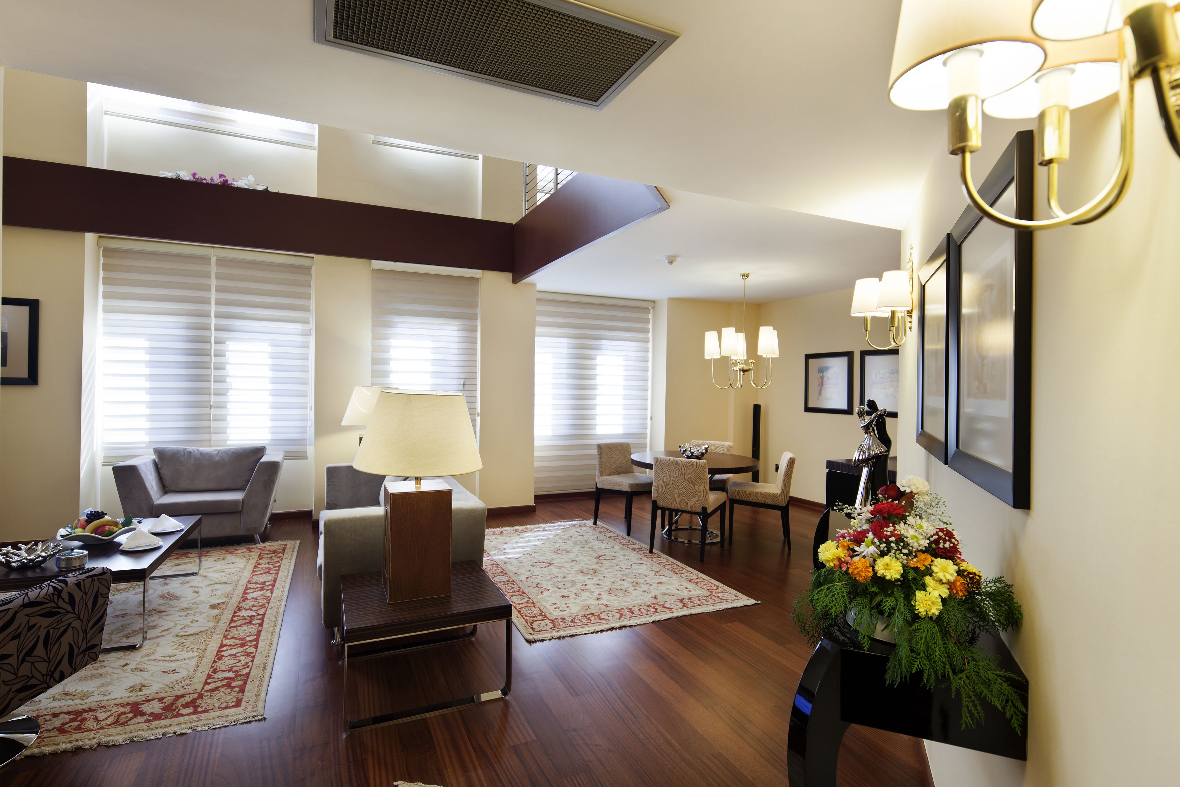 A spacious living room with a sofa, coffee table, and a dining area with chairs. There is a large window with blinds, a fireplace, and a wall-mounted lamp.